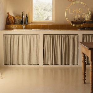 Op de afbeelding: Een keuken met witte kasten en een beige betegelde achterwand. De kasten zijn bedekt met beige stoffen gordijnen. Op de voorgrond staat een houten tafel met gedraaide poten.