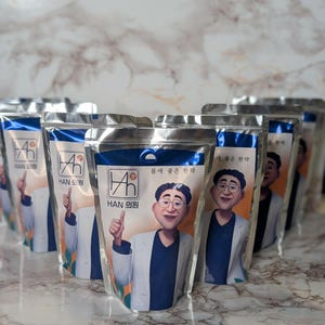 May include: Several silver pouches with a blue top, featuring a cartoon illustration of a man in a white coat and glasses giving a thumbs up. The text "HAN 의원" is visible.