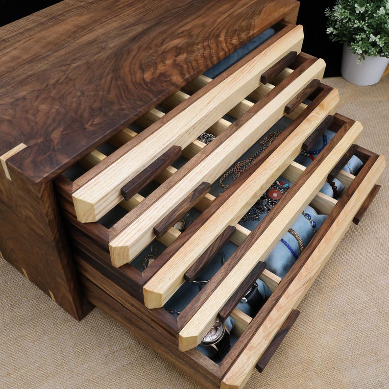 Puede incluir: Una caja de madera para joyas con varios cajones. Los cajones est&aacute;n hechos de madera clara y oscura y tienen un forro de terciopelo azul. La caja est&aacute; abierta y los cajones est&aacute;n sacados, revelando el interior.