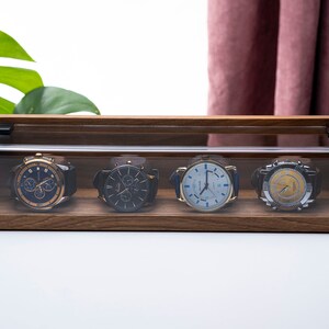 Puede incluir: Una caja de madera para relojes con una tapa de acr&iacute;lico transparente, que muestra cuatro relojes de pulsera con diferentes caras y correas de colores.