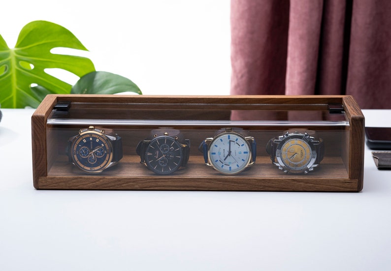 Puede incluir: Una caja de madera para relojes con una tapa de acr&iacute;lico transparente, que muestra cuatro relojes de pulsera con diferentes caras y correas de colores.