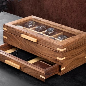 May include: A wooden watch box with a glass top and a drawer. The box has a light brown finish and is made of wood with a natural grain. The drawer is open and shows a black leather watch case inside.