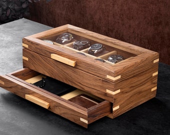 May include: A wooden watch box with a glass top and a drawer. The box has a light brown finish and is made of wood with a natural grain. The drawer is open and shows a black leather watch case inside.