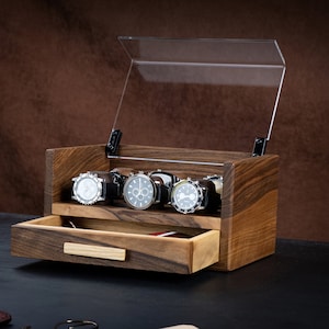 Puede incluir: Una caja expositora de madera rectangular con una tapa abatible de acrílico transparente. El interior cuenta con un estante escalonado que sostiene tres relojes de pulsera y un cajón inferior con un tirador de madera clara para guardar objetos.