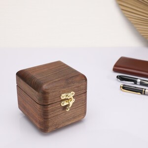 May include: A small, square wooden box with a hinged lid and a gold latch. The box is made of dark wood with a natural grain pattern.