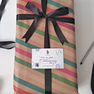 Puede incluir: Una caja de regalo de rayas marrones y verdes con una cinta negra atada en un lazo. Una tarjeta blanca con texto negro dice "Estoy tan contento de que existas. Feliz cumplea&ntilde;os Mary J."