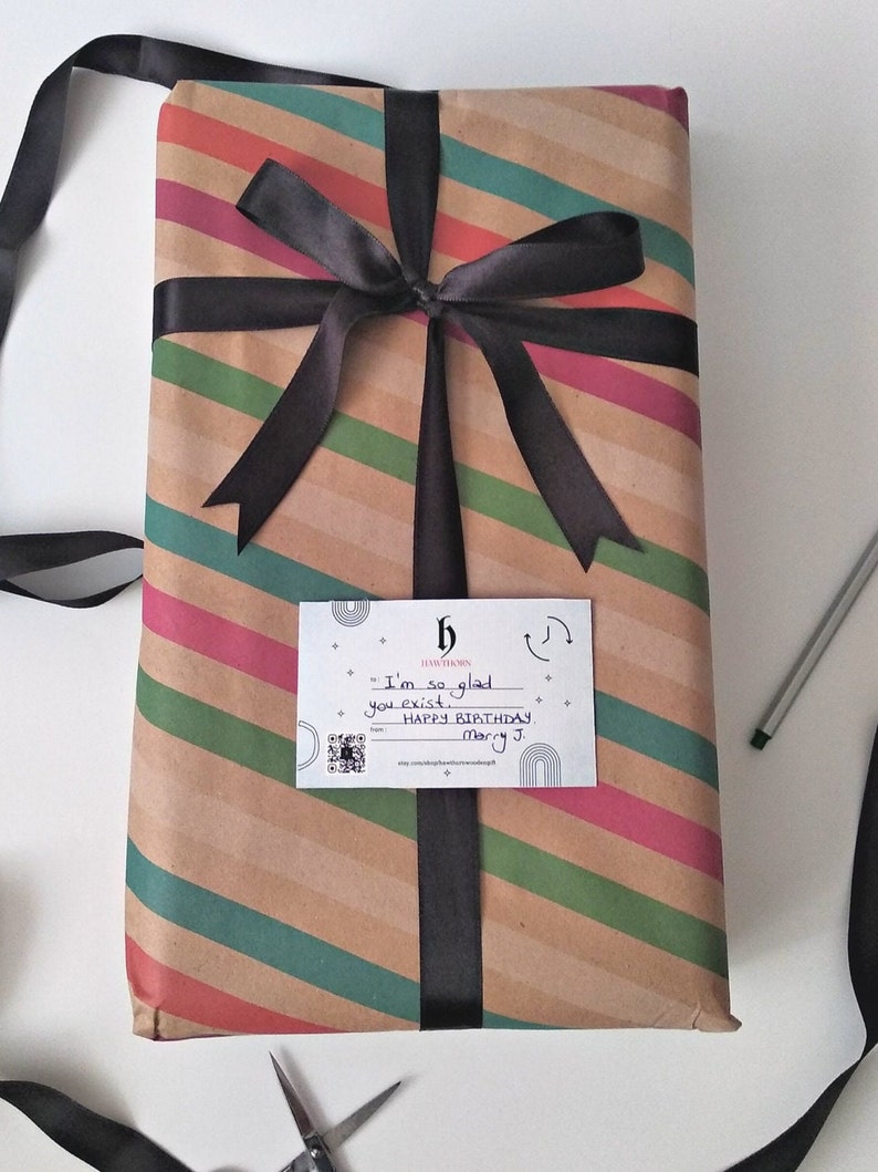 Puede incluir: Una caja de regalo de rayas marrones y verdes con una cinta negra atada en un lazo. Una tarjeta blanca con texto negro dice "Estoy tan contento de que existas. Feliz cumplea&ntilde;os Mary J."
