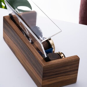 Puede incluir: Una caja de madera para relojes con una tapa de acr&iacute;lico transparente. La caja tiene una veta de madera oscura y est&aacute; abierta para revelar varios relojes en su interior.