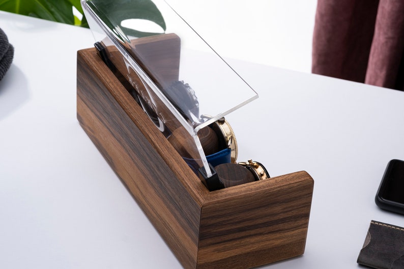 Puede incluir: Una caja de madera para relojes con una tapa de acr&iacute;lico transparente. La caja tiene una veta de madera oscura y est&aacute; abierta para revelar varios relojes en su interior.