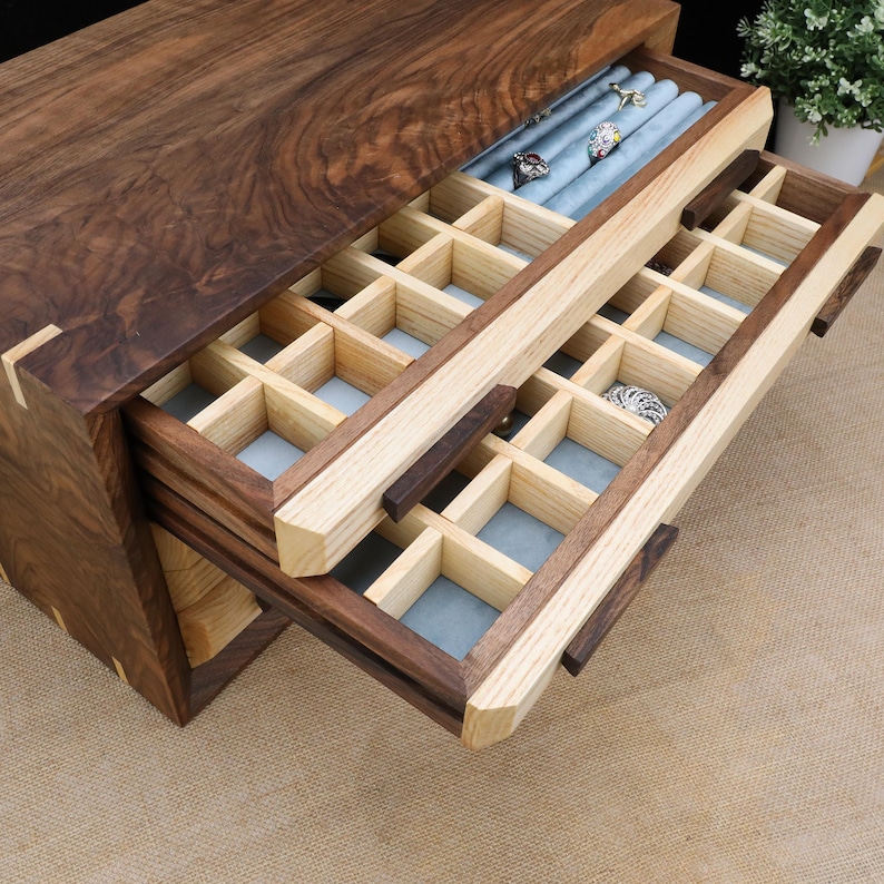 Puede incluir: Una caja de madera para joyas con dos cajones. El caj&oacute;n superior est&aacute; abierto y tiene un forro de terciopelo azul con compartimentos para anillos. El caj&oacute;n inferior tambi&eacute;n est&aacute; abierto y tiene un interior de madera clara con compartimentos para joyas.