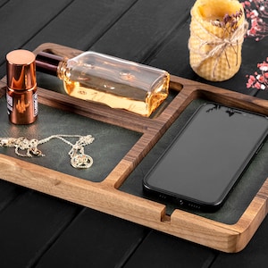 May include: A wooden tray with four compartments, featuring a gold necklace, a clear glass bottle with a brown cap, a copper-coloured bottle, and a black smartphone. The tray is on a black wooden surface.