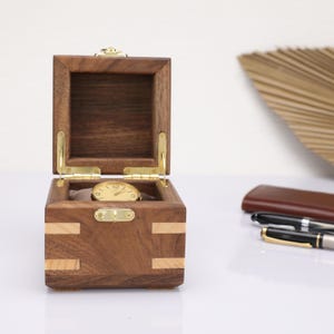 May include: A wooden watch box with a hinged lid and a gold-coloured watch inside. The box is made of dark wood with light wood accents.