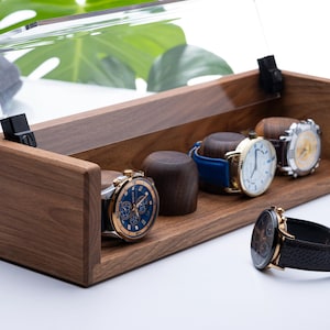 May include: A wooden watch display case with a clear acrylic lid, showcasing several wristwatches with various band and face designs. The watches include gold, silver, and blue accents. The case is made of dark wood.
