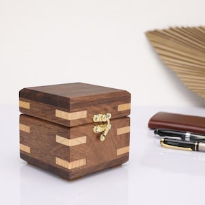 May include: A small wooden box with a hinged lid and a brass latch. The box is made of dark wood with light wood accents.