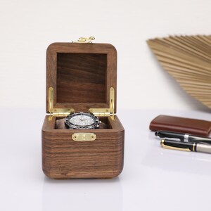 May include: A dark brown wooden watch box with a hinged lid and brass accents. The box is open and a silver watch is inside.