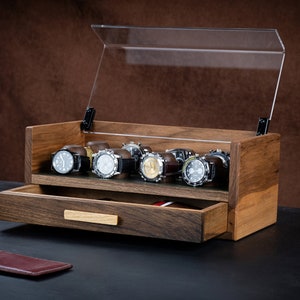 Op de afbeelding: Een houten horlogebox met een doorzichtig deksel en een lade. De doos heeft acht vakken voor horloges en is gemaakt van donker hout. De lade is open en is ook gemaakt van donker hout.