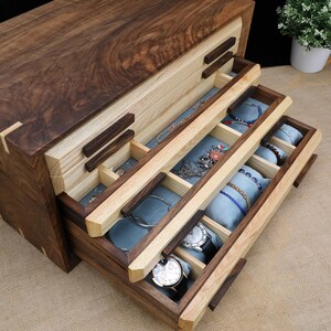 Puede incluir: Una caja de joyer&iacute;a de madera con tres cajones. Los cajones est&aacute;n forrados con terciopelo azul y tienen compartimentos para guardar joyas. La caja est&aacute; hecha de madera de nogal y arce.