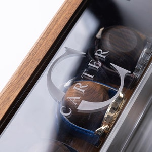 Puede incluir: Una caja de madera para relojes con tapa de cristal. La caja tiene una inscripci&oacute;n personalizada que dice "CARTER". La caja est&aacute; abierta y hay dos relojes dentro.