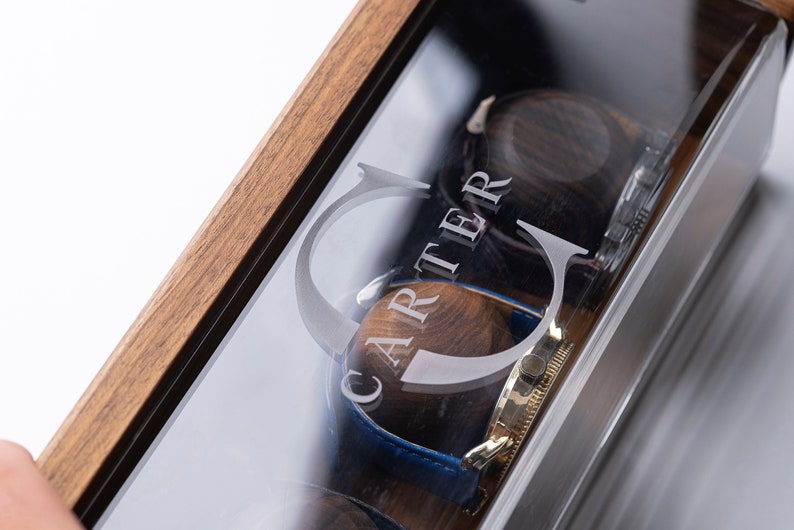 Puede incluir: Una caja de madera para relojes con tapa de cristal. La caja tiene una inscripci&oacute;n personalizada que dice "CARTER". La caja est&aacute; abierta y hay dos relojes dentro.