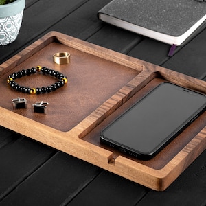 Peut inclure: Un plateau en bois avec deux compartiments. Le plus grand compartiment contient un bracelet en perles noires, une bague en or et deux boutons de manchette en argent. Le plus petit compartiment contient un smartphone noir.