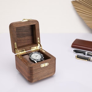 May include: A wooden watch box with a hinged lid and a brass latch. The box is open and a silver watch is inside.