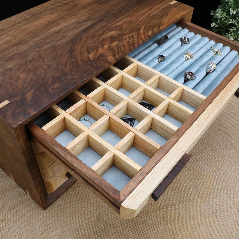 Puede incluir: Una caja de joyer&iacute;a de madera con un caj&oacute;n que se abre para revelar un compartimento forrado de terciopelo azul con m&uacute;ltiples ranuras para anillos y aretes. El caj&oacute;n tambi&eacute;n tiene una fila de rollos forrados de terciopelo azul para guardar anillos.