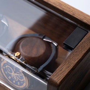 Puede incluir: Una caja de madera para relojes con tapa de cristal transparente. La caja tiene un tinte marr&oacute;n oscuro y est&aacute; dise&ntilde;ada para guardar dos relojes. La caja tiene un cierre negro en el lateral.