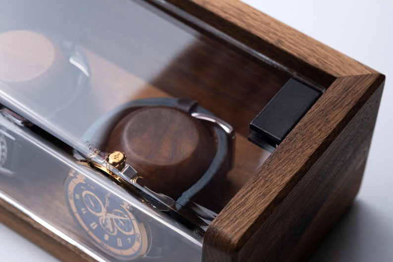 Puede incluir: Una caja de madera para relojes con tapa de cristal transparente. La caja tiene un tinte marr&oacute;n oscuro y est&aacute; dise&ntilde;ada para guardar dos relojes. La caja tiene un cierre negro en el lateral.