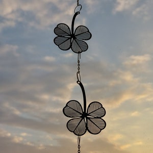 May include: A decorative hanging ornament featuring two black metal four-leaf clovers connected by a chain, with a faceted crystal ball at the bottom. The ornament hangs against a sunset sky, with a cityscape visible in the distance.