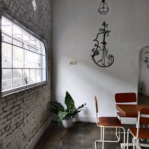 May include: A white brick wall with a window and a plant in a white pot. There is a black metal sculpture of a violin on the wall. Two white chairs with wooden seats are in front of a wooden table with a white top.