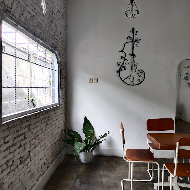 May include: A white brick wall with a window and a plant in a white pot. There is a black metal sculpture of a violin on the wall. Two white chairs with wooden seats are in front of a wooden table with a white top.