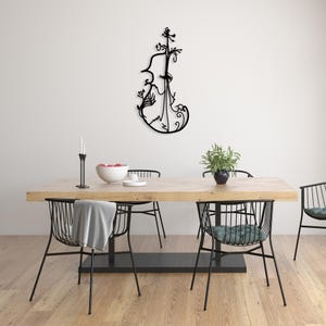 May include: A black metal wall sculpture of a violin with intricate floral details. The sculpture is hanging on a white wall above a wooden dining table with black metal chairs.
