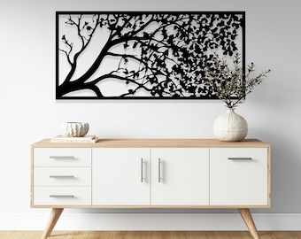 May include: A black metal tree wall art decor with a detailed silhouette of branches and leaves. The artwork is mounted on a white wall above a white and wood-tone cabinet with silver handles and legs. A vase with greenery is on the cabinet.