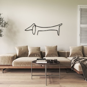 May include: A black line drawing of a dachshund on a white wall above a beige sectional sofa with three pillows. A wooden coffee table with a black book and two black coasters sits in front of the sofa.