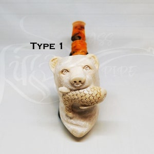 May include: A carved tobacco pipe featuring a bear holding a fish. The bear is cream-colored with detailed features. The stem is orange and black. The text "TYPE 1" is above the bear's head.
