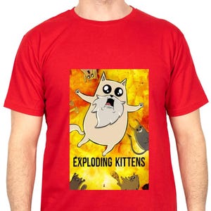 Peut inclure: T-shirt rouge avec un dessin animé du jeu Exploding Kittens. Le graphisme montre un chat brun clair avec une barbe blanche et de grands yeux sur un fond jaune et orange. Les mots "EXPLODING KITTENS" sont imprimés en dessous.