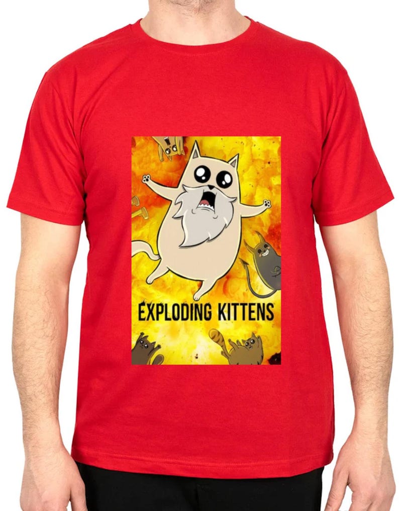 May include: Red t-shirt featuring a cartoon image from the game Exploding Kittens. The graphic shows a light brown cat with a white beard and wide eyes against a yellow and orange background. The words "EXPLODING KITTENS" are printed below the image.