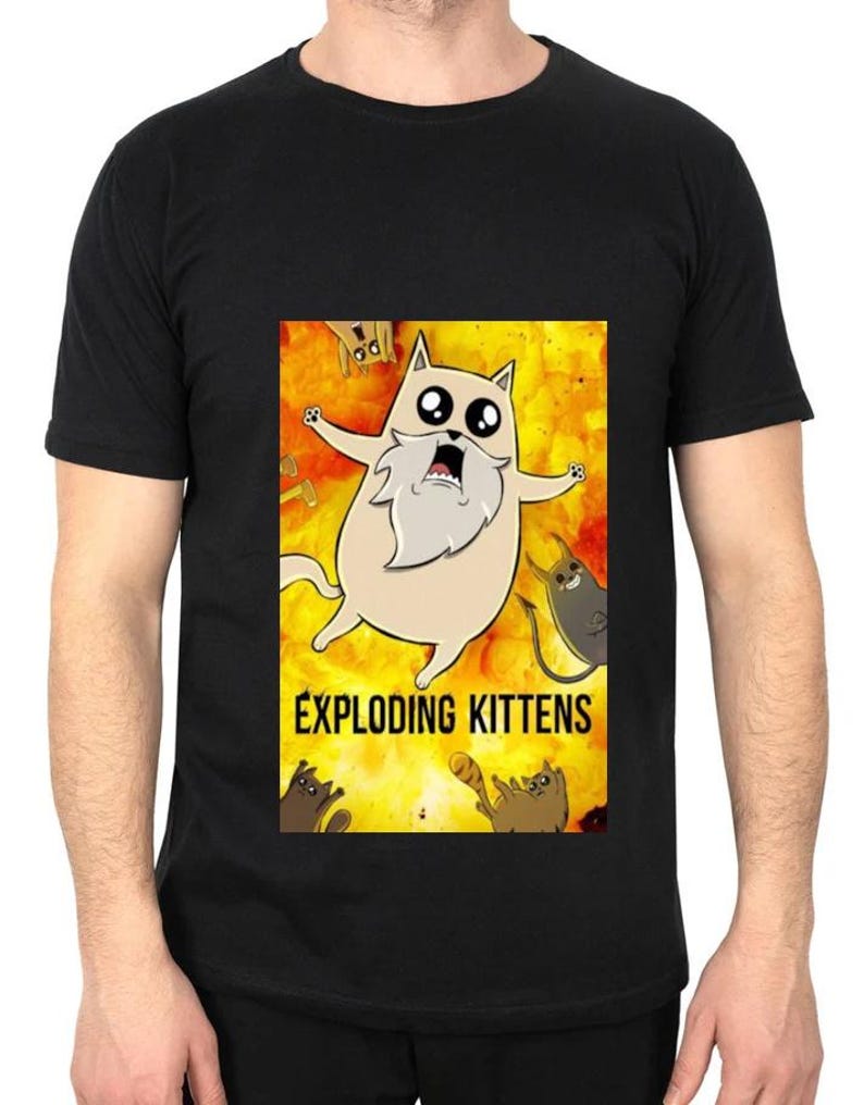 May include: Black t-shirt featuring a colorful graphic from the card game Exploding Kittens. The design includes a cartoon cat with a long white beard and other cartoon cats on a yellow and orange background.