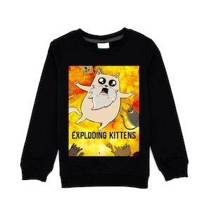 Peut inclure: Sweat-shirt noir à manches longues avec un dessin animé du jeu Exploding Kittens. Le dessin animé représente un chat surpris, d'autres chats et le titre du jeu sur un fond jaune et orange.
