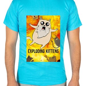 May include: A light blue t-shirt featuring a cartoon image from the game "Exploding Kittens." The graphic shows a surprised cat with a long white beard against a fiery background, with the game's title below.