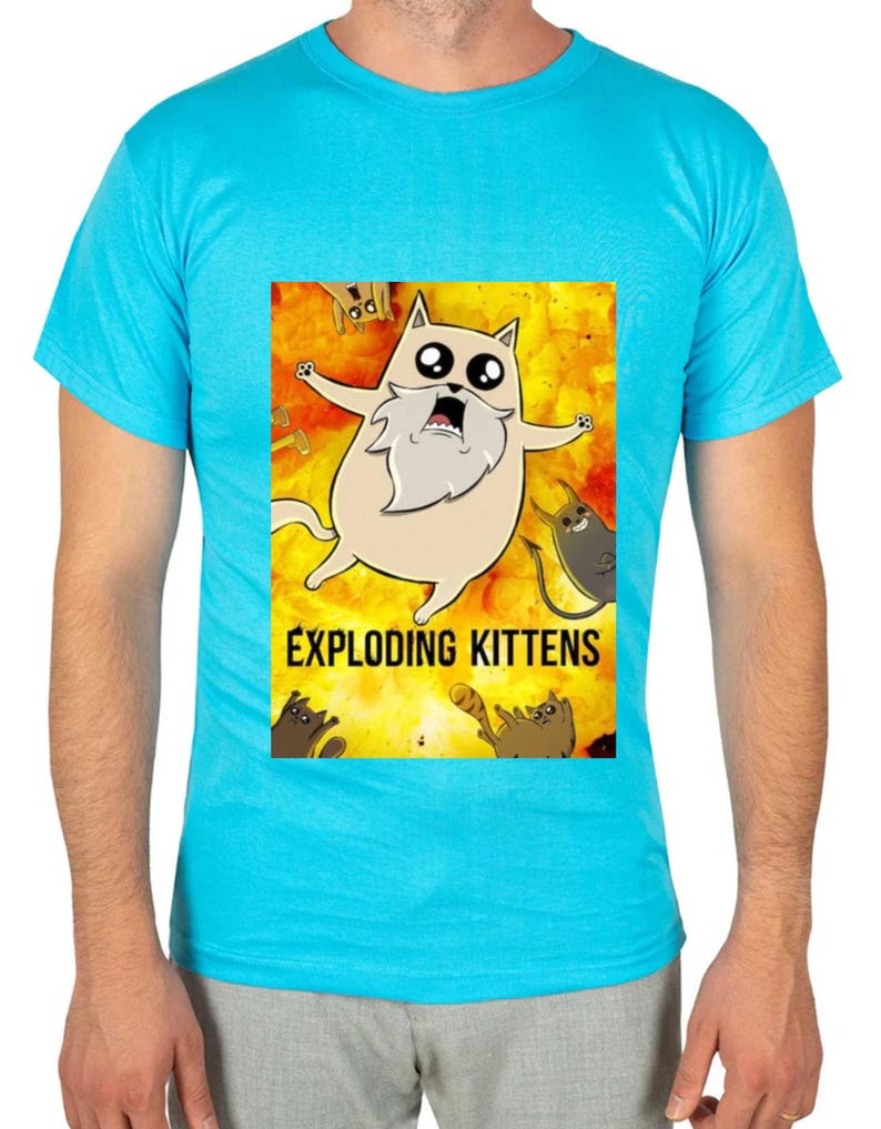 May include: A light blue t-shirt featuring a cartoon image from the game "Exploding Kittens." The graphic shows a surprised cat with a long white beard against a fiery background, with the game's title below.