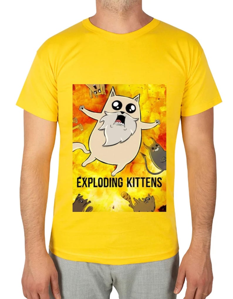 May include: Yellow t-shirt featuring a cartoon image of a cat with a long white beard and the text "EXPLODING KITTENS". The cat is surrounded by other cartoon cats on a yellow and orange background.