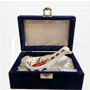 May include: A white ceramic spoon rest with a red chilli pepper design, accented with green leaves and blue dots, inside a navy blue velvet box with a gold clasp. A kitchen accessory.