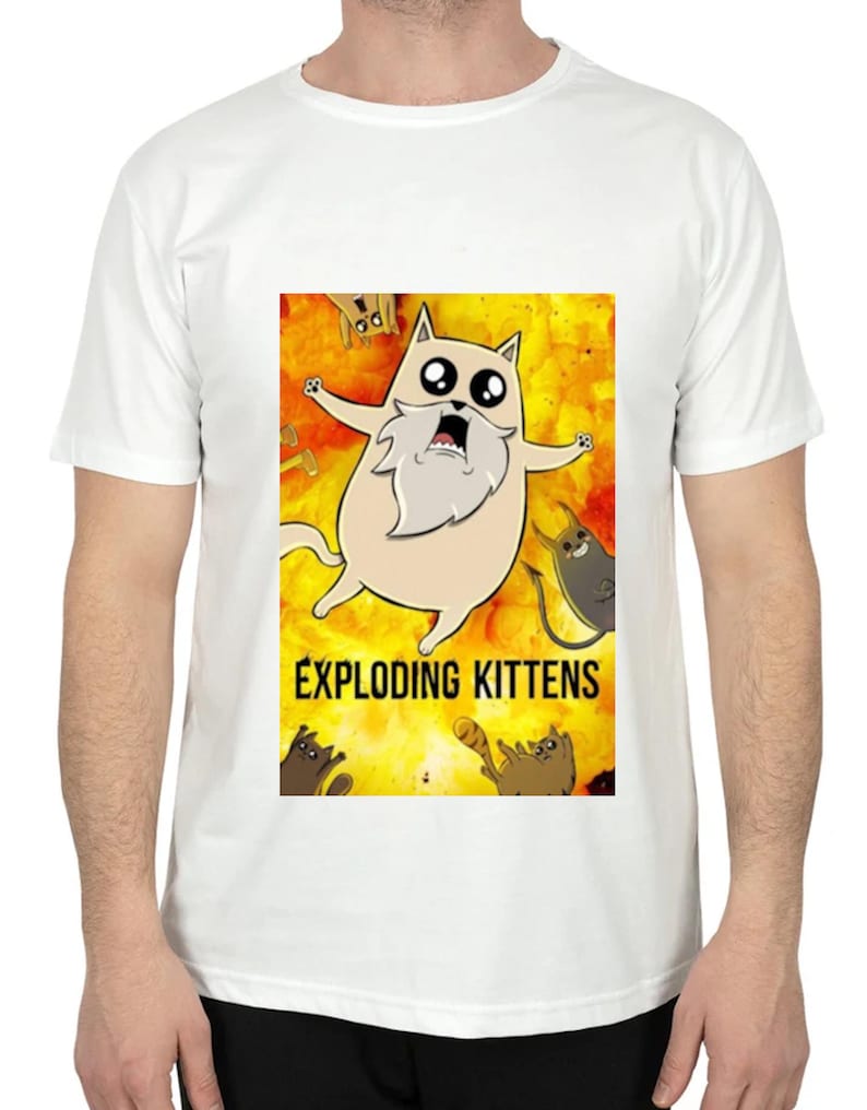 May include: White t-shirt featuring a cartoon graphic from the game Exploding Kittens. The graphic shows a surprised cat with a long white beard against a yellow and orange explosion background. The words "EXPLODING KITTENS" are printed below the cat.