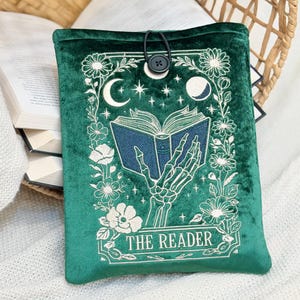Può includere: Custodia per libri in velluto verde smeraldo con un design ricamato. Il design presenta una mano scheletrica che tiene un libro aperto, circondata da fiori, lune e stelle. Il testo "THE READER" è in basso. Chiusura con bottone e asola nera.