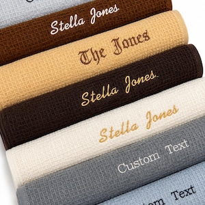 Könnte beinhalten: Ein Stapel gerollter Waffelhandtücher in verschiedenen Farben, darunter Grau, Braun und Beige. Einige Handtücher sind mit den Texten "Stella Jones", "The Jones" und "Custom Text" bestickt. Die Handtücher haben eine strukturierte Oberfläche.