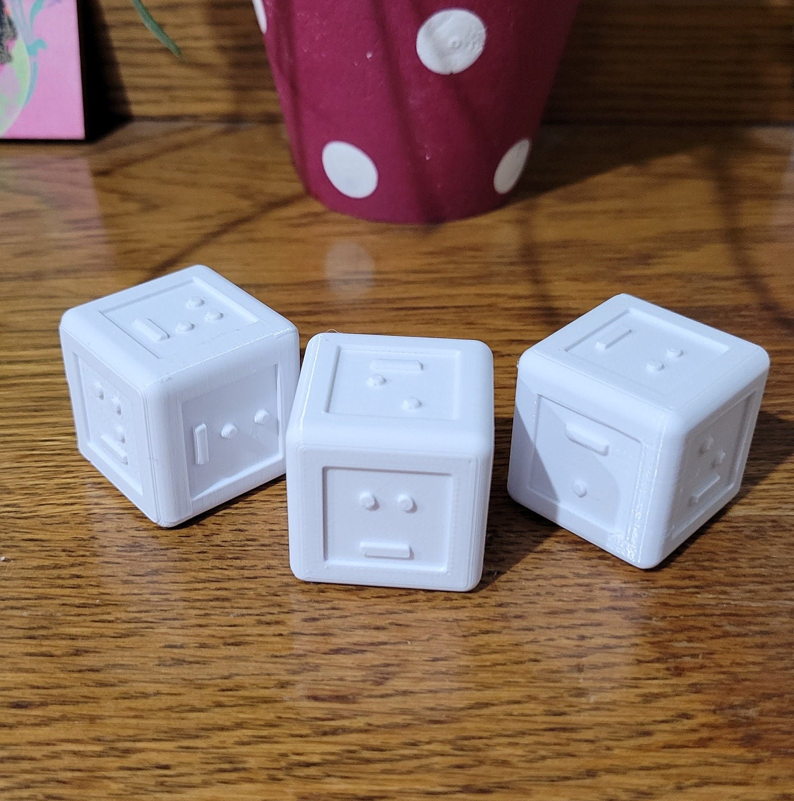 Three white cube dice on a wooden table, each face showing raised Braille dots with a short bar beneath to mark which way is up.