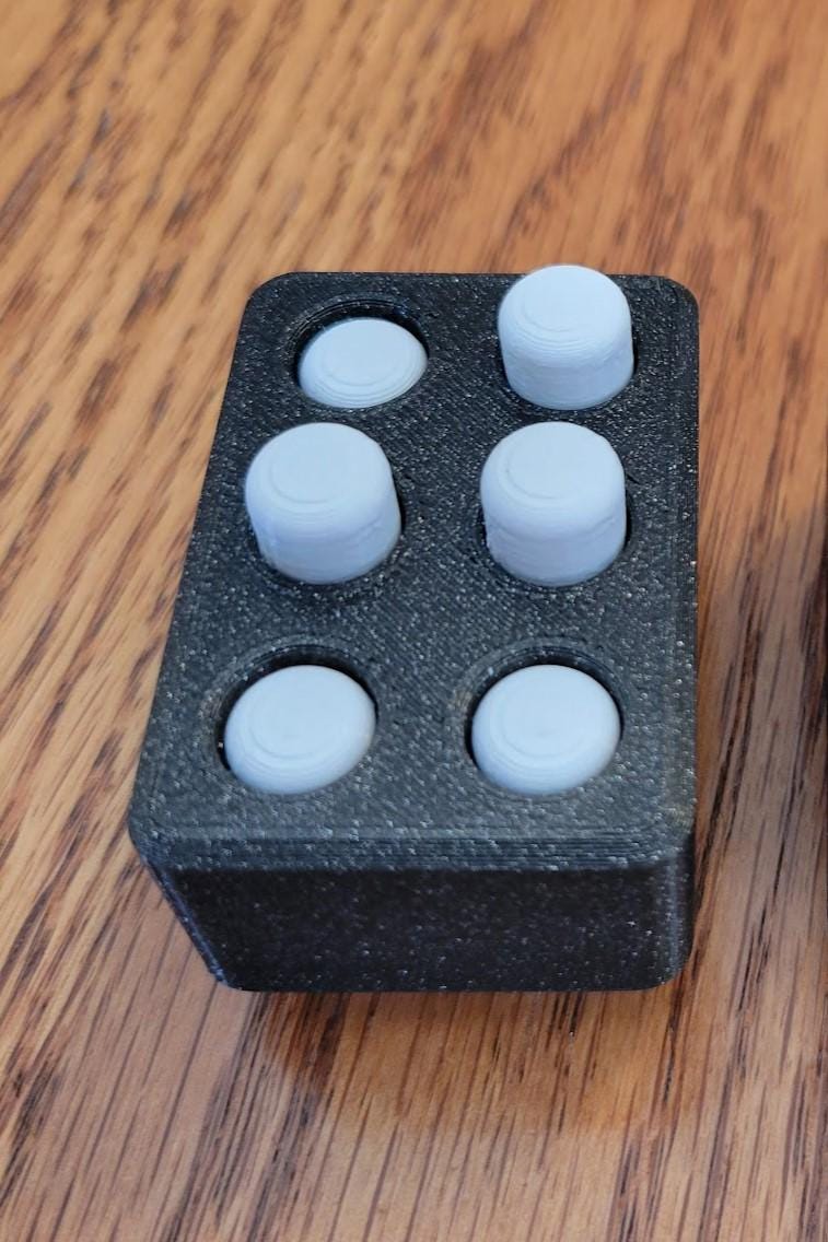 Black rectangular fidget with six light blue round buttons in the 2-by-3 layout of a Braille cell, some raised and some pressed down.