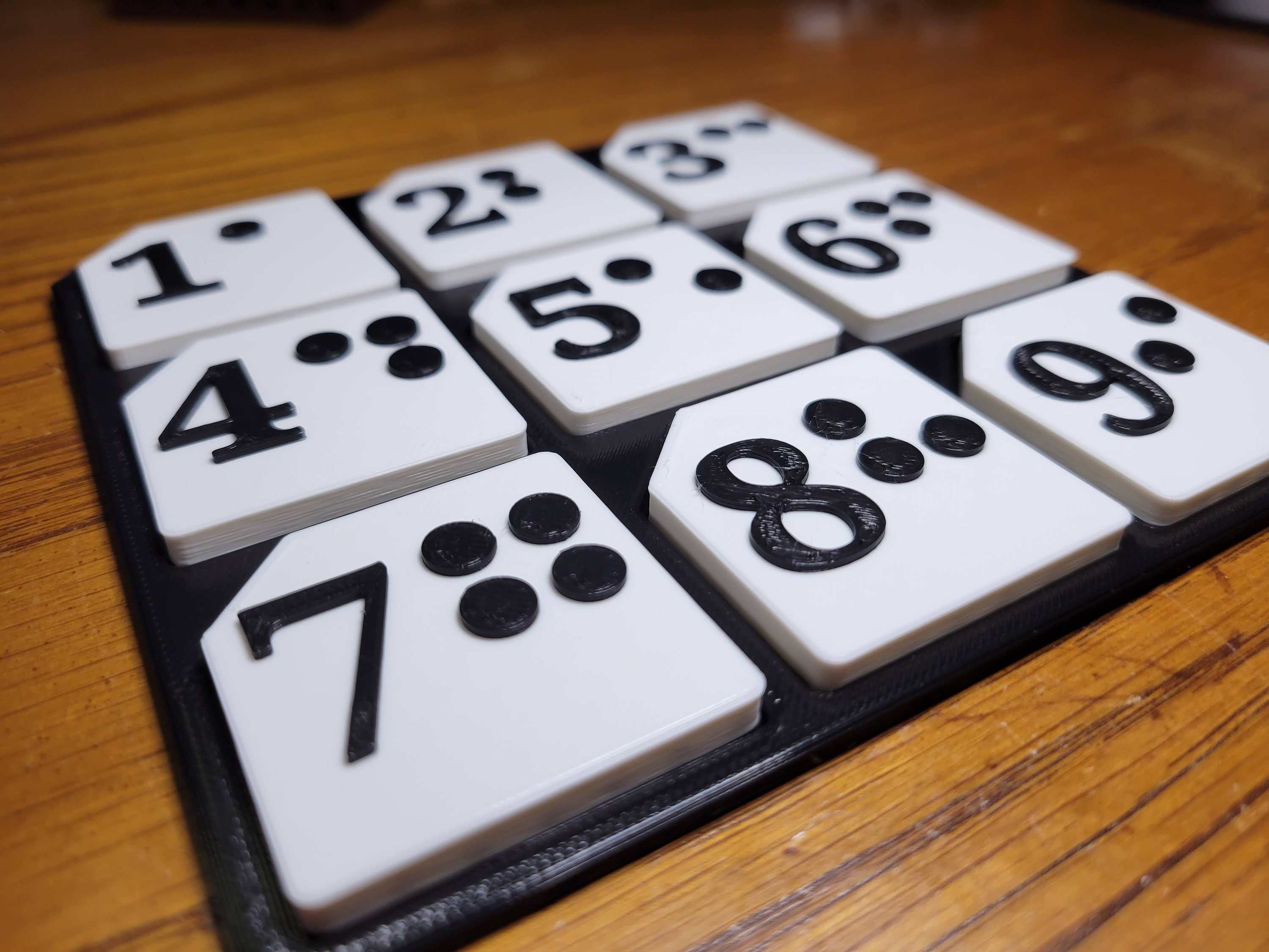 Close-up of the 3-by-3 board in the both style: white tiles, each with a raised black number and its Braille dots.