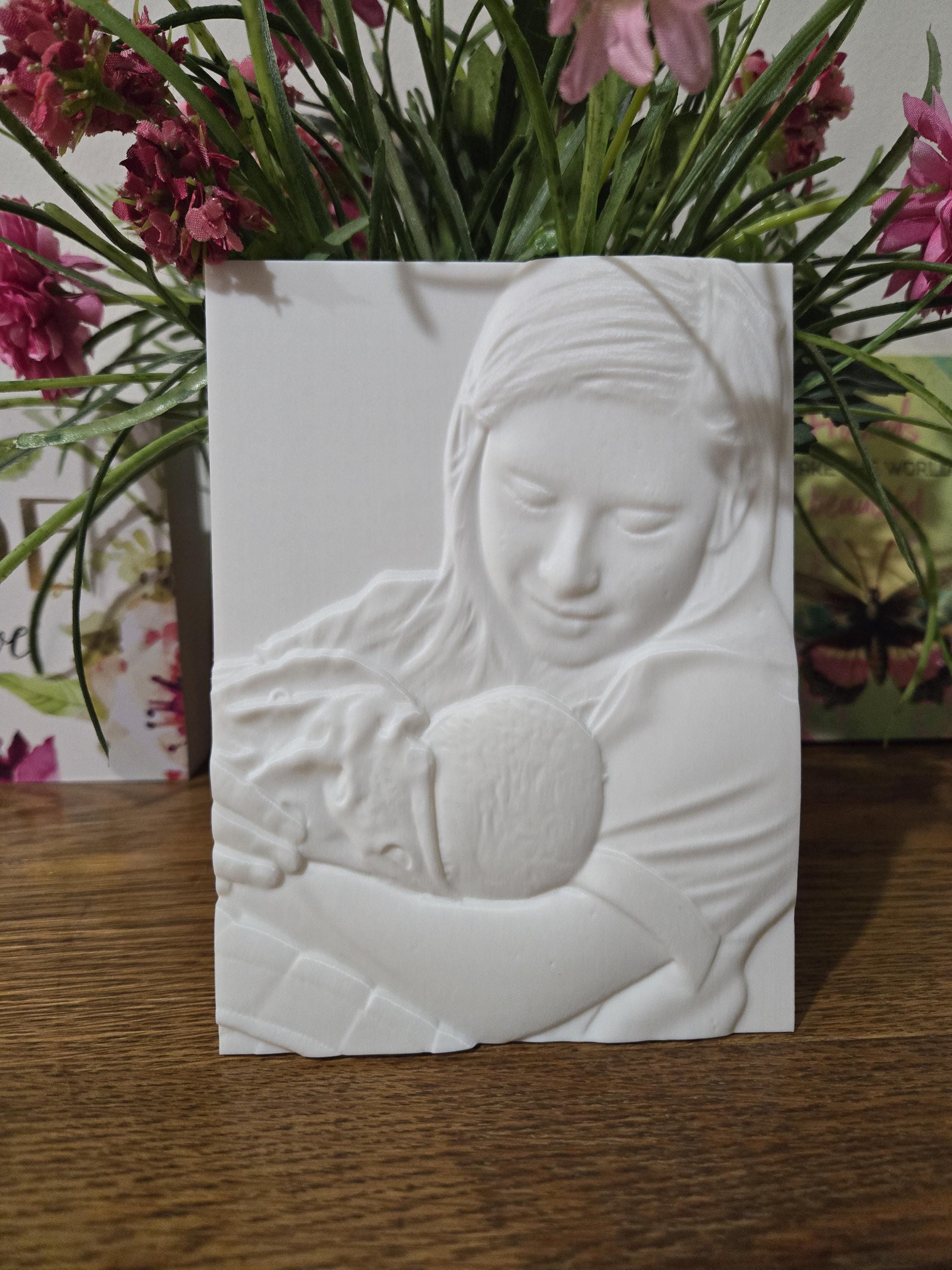 White tactile picture standing in front of flowers: a raised relief of a woman holding a baby close.